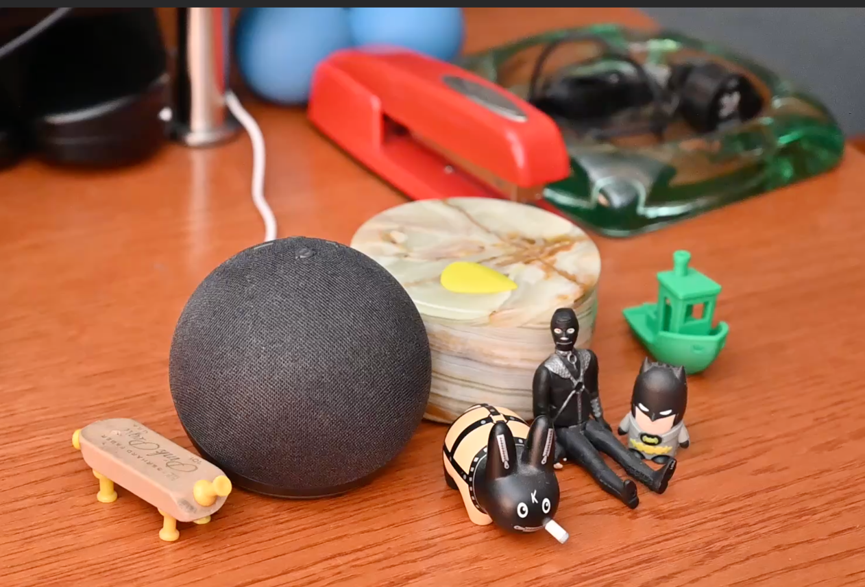 Black Amazon Echo Dot™ on a wooden desk, surrounded by a Bondage Labbit™, an action figure of The Gimp™, a Batman™ USB thumb drive, a green 3D-printed Benchy boat, a Little Pink Pig™, and a red Swingline™ stapler.